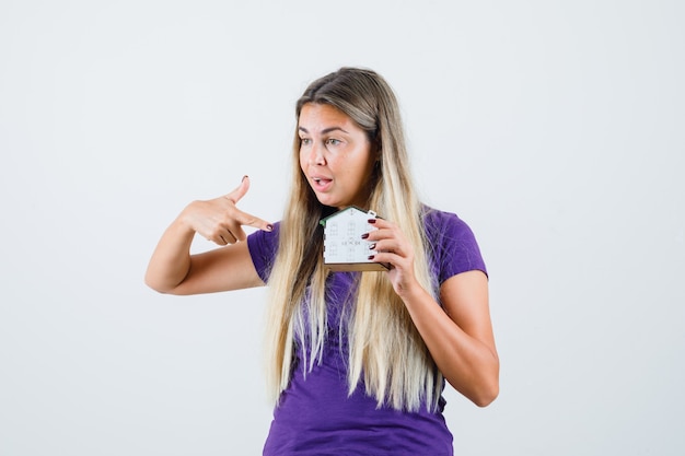 Senhora loira em t-shirt violeta, apontando para o modelo da casa e olhando perplexa, vista frontal.