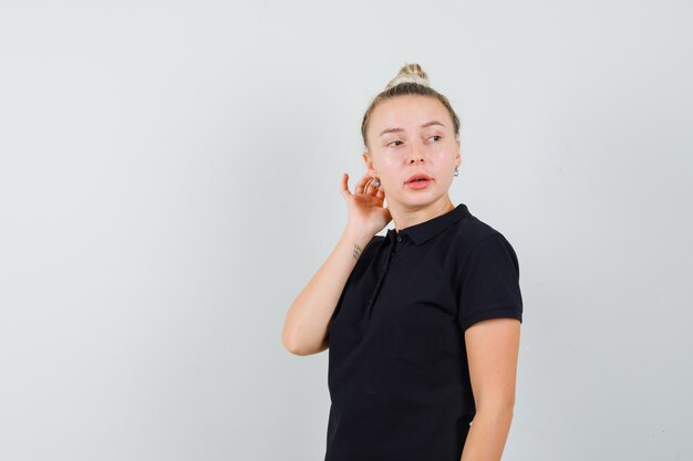 Senhora loira em uma camiseta preta olhando por cima do ombro e parecendo curiosa, vista frontal.