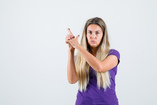 Senhora loira mostrando gesto de arma em t-shirt violeta e olhando confiante, vista frontal.