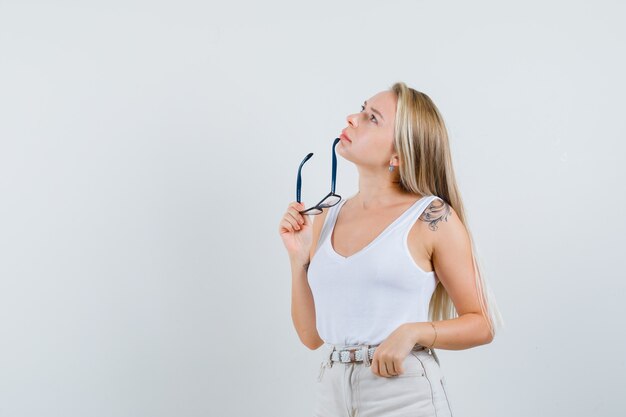 Senhora loira segurando óculos na camiseta, calça e olhando pensativa, vista frontal.