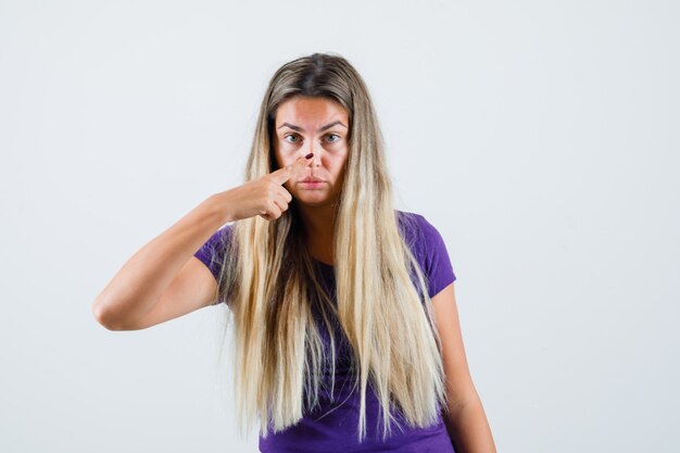 Senhora loira tocando o nariz com o dedo na t-shirt violeta, vista frontal.