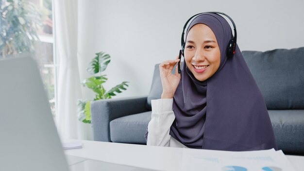 senhora muçulmana usar fone de ouvido usando laptop para falar com colegas sobre o plano de videoconferência enquanto trabalha em casa na sala de estar.