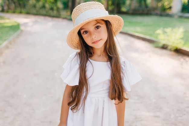 Foto grátis senhora na moda com cílios longos, olhando com interesse em pé na estrada com chapéu elegante. retrato ao ar livre da tímida garota de cabelos castanhos usando um lindo vestido branco.