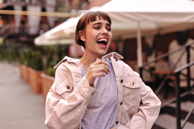 Senhora otimista com penteado curto em roupa leve rindo na rua Mulher morena alegre com jaqueta rosa jeans posando do lado de fora