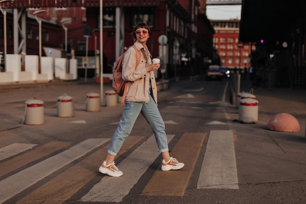 Senhora otimista com roupa leve sai garota moderna em óculos de tênis jeans e jaqueta bege com mochila segura xícara de chá na rua