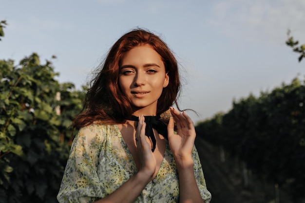 Senhora ruiva com curativo preto no pescoço no elegante vestido amarelo e verde sorrindo e olhando para a câmera posando no fundo das vinhas