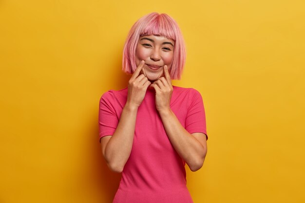 Senhora sensual e positiva com cabelo rosa, mantém os dedos perto dos cantos da boca, força o sorriso, finge estar de bom humor