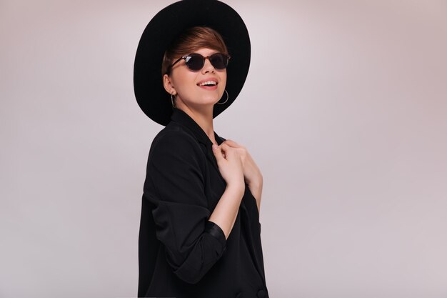 Senhora sorridente de óculos e chapéu preto, posando em fundo branco. Mulher alegre em uma jaqueta preta sorrindo em um cenário isolado