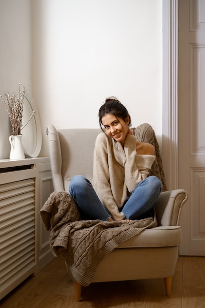 Senhora sorridente em roupas da moda inteligente está sentada na poltrona. sorrindo, sentado em uma atmosfera relaxante dentro de casa