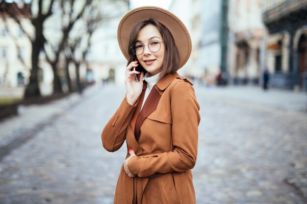 Senhora sorridente no chapéu largo, falando no smartphone outono rua