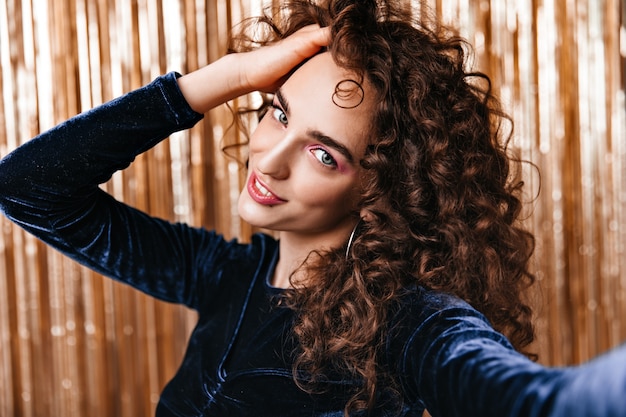 Senhora tocando seu cabelo encaracolado e tirando uma selfie sobre fundo dourado