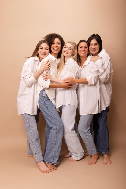 Senhoras de diversas idades felizes de comprimento total em camisas brancas e jeans sorrindo para a câmera contra o fundo bege