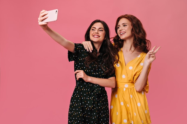 Senhoras encaracoladas em elegantes vestidos de verão tiram selfie em fundo rosa Lindas namoradas em macacões brilhantes posando para foto