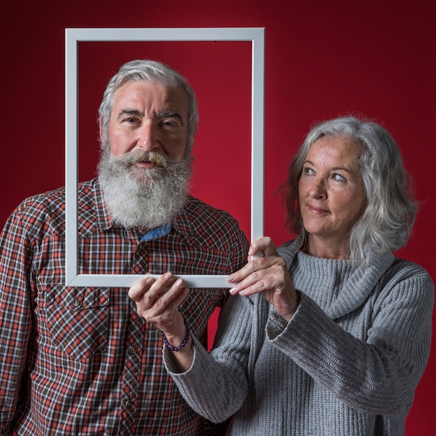 Sênior mulher segurando a borda do quadro branco na frente do rosto do marido contra o pano de fundo vermelho