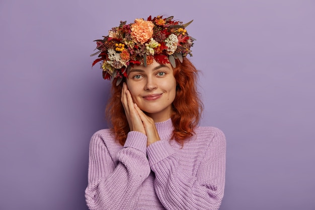 Foto grátis sensual senhora europeia ruiva mantém ambas as mãos perto das bochechas, sorri com covinhas nas bochechas, usa uma bela grinalda de outono e veste um macacão de malha de tamanho grande