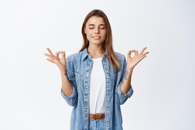 Sentindo zen no corpo. Mulher jovem relaxada, calma e tranquila em pose de meditação, fecha os olhos e respira, em pé sobre o branco