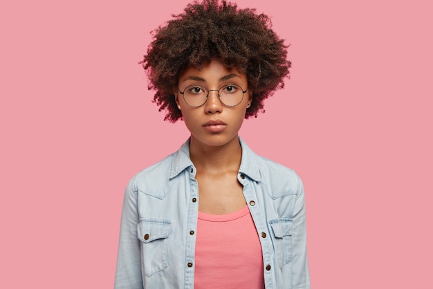 Séria adorável mulher afro-americana com cabelo encaracolado, vestida com uma jaqueta jeans da moda, posa contra a parede rosa, ouve as informações necessárias. conceito de pessoas, beleza e etnia