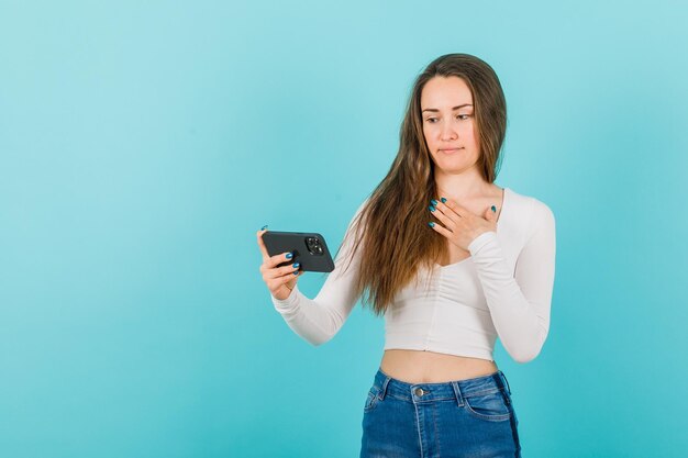 Sério, garota está olhando para a tela do smartphone segurando a mão no peito no fundo azul