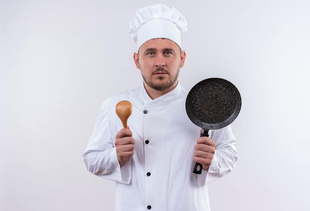 Sério, jovem e bonito cozinheiro em uniforme de chef segurando uma colher e uma frigideira olhando para um espaço em branco isolado