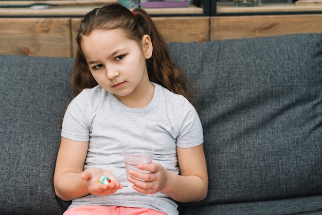 Sério menina sentada no sofá segurando pílulas e copo de água na mão