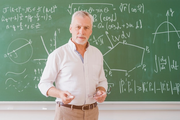 Sério professor do sexo masculino em pé no quadro-negro com gráfico e equação e olhando para a câmera