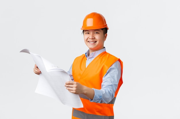 Setor de construção e conceito de trabalhadores industriais. Arquiteto asiático sorridente e confiante, engenheiro-chefe com capacete e jaqueta reflexiva segurando plantas, inspecionando a empresa, parede branca