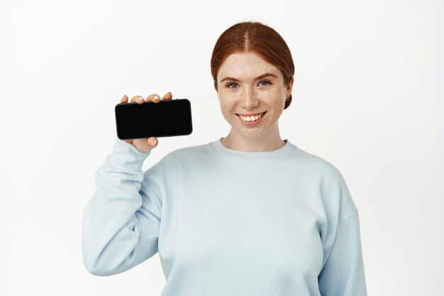 Seu aplicativo de smartphone. Sorridente jovem ruiva mostrando a tela do celular em branco.