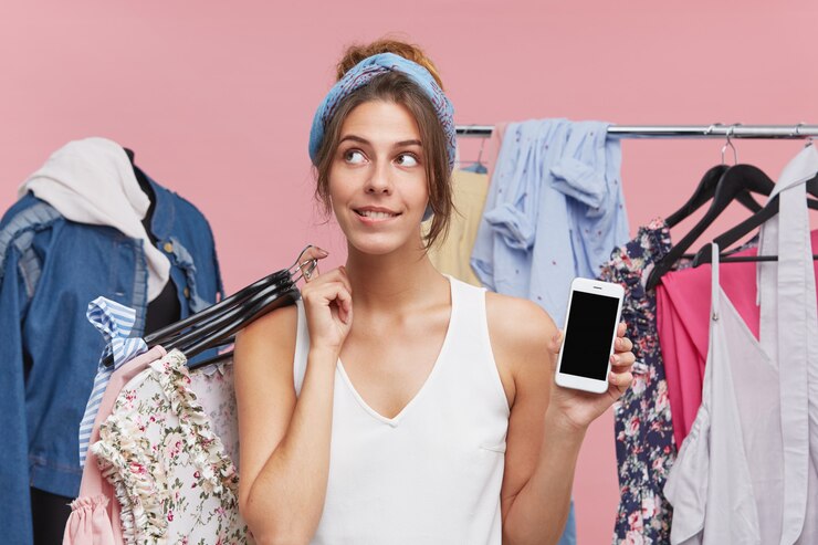 Foto grátis shopaholic feminino em pé na boutique, levando muitos cabides com roupas para experimentar, olhando com uma expressão sonhadora de lado, decidindo o que levar, mantendo o celular moderno por outro lado