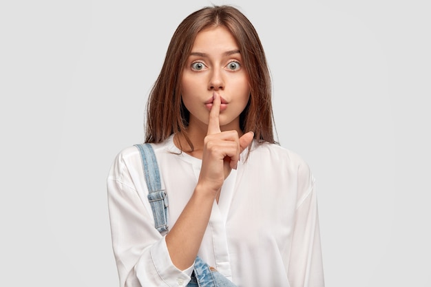 Foto grátis shush, fique em silêncio! mulher adorável e séria pede para manter as informações secretas confidenciais, vestida com uma camisa branca enorme e um macacão jeans, posa contra uma parede branca. conceito de conspiração