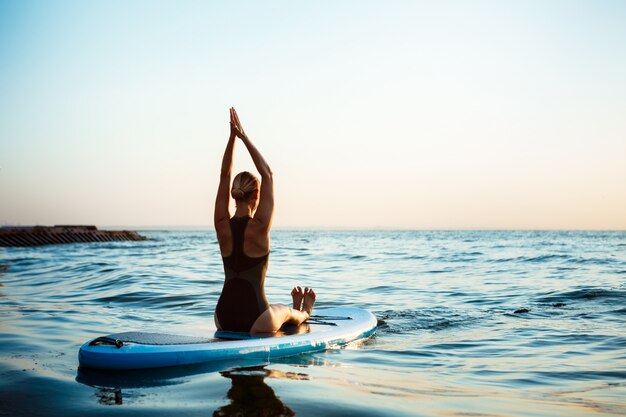 Silhueta de mulher bonita praticando ioga na prancha de surf ao nascer do sol.