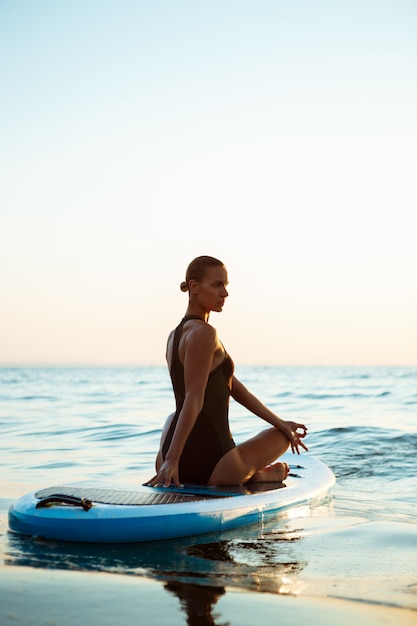 Silhueta de mulher bonita praticando ioga na prancha de surf ao nascer do sol.