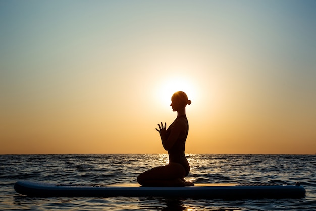 Foto grátis silhueta de mulher bonita praticando ioga na prancha de surf ao nascer do sol.