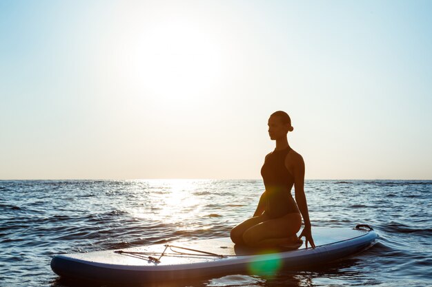 Silhueta de mulher bonita praticando ioga na prancha de surf ao nascer do sol.