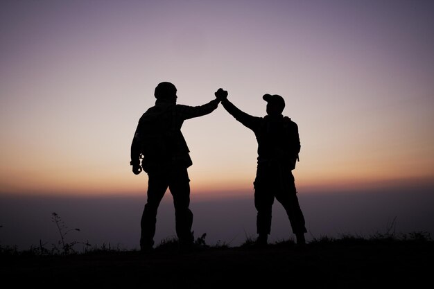 Silhueta de trabalho em equipe, ajudando a confiar na mão, ajudando o sucesso nas montanhas Os caminhantes comemoram com as mãos para cima Ajudam uns aos outros no topo da montanha e da paisagem do pôr do sol