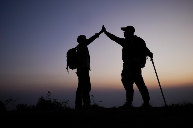 Silhueta de trabalho em equipe, ajudando a confiar na mão, ajudando o sucesso nas montanhas Os caminhantes comemoram com as mãos para cima Ajudam uns aos outros no topo da montanha e da paisagem do pôr do sol