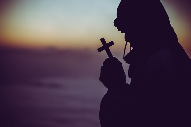 Silhueta de um homem rezando com uma cruz na mão ao nascer do sol.