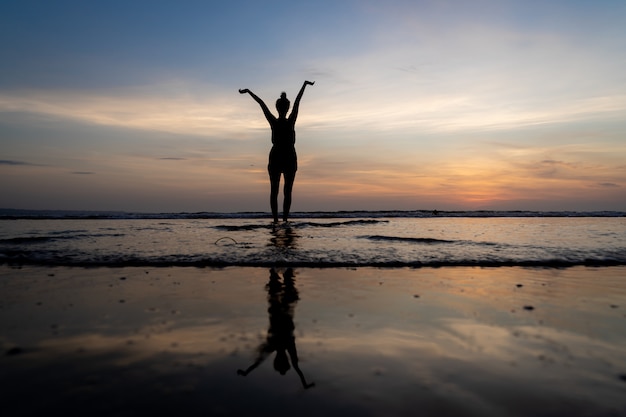 Silhueta de uma garota de pé na água com os braços levantados e seu reflexo na água