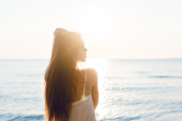 Silhueta de uma garota magro em uma praia com o sol poente. Ela usa um vestido branco. Ela tem cabelo comprido que voa no ar. Seus braços esticados no ar