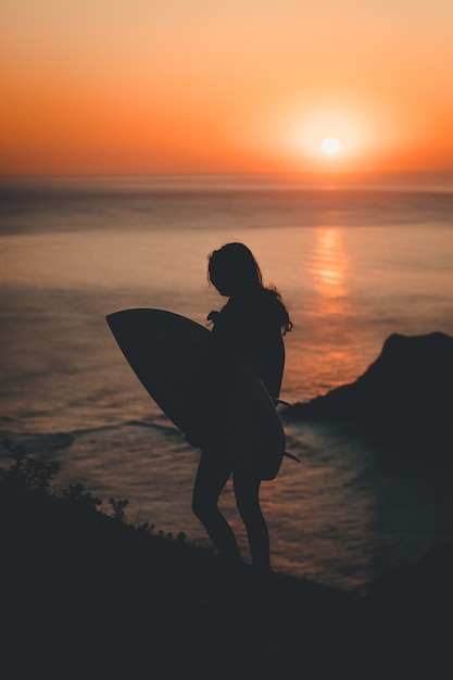 Silhueta de uma mulher solitária segurando uma prancha de surf, caminhando à beira-mar ao pôr do sol