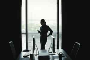 Foto grátis silhueta escura da mulher em pé contra a janela no escritório e usando o smartphone