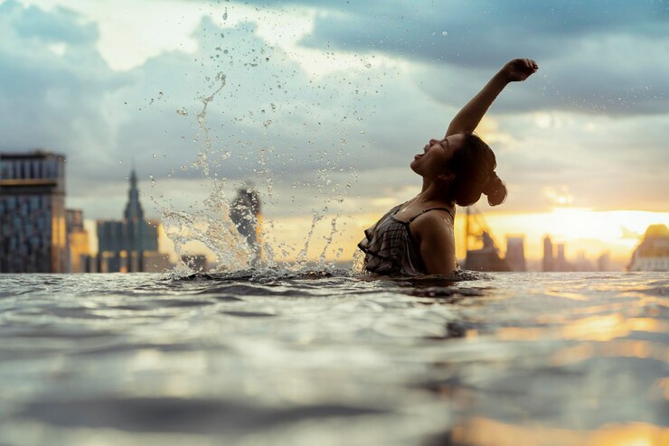 Foto grátis silhueta negra de mulher asiática respingo de água nas férias de verão relaxando na piscina infinita com vista para o pôr do sol do mar azul com arranha-céus skyscape urbano centro da cidade estilo de vida saudável felicidade