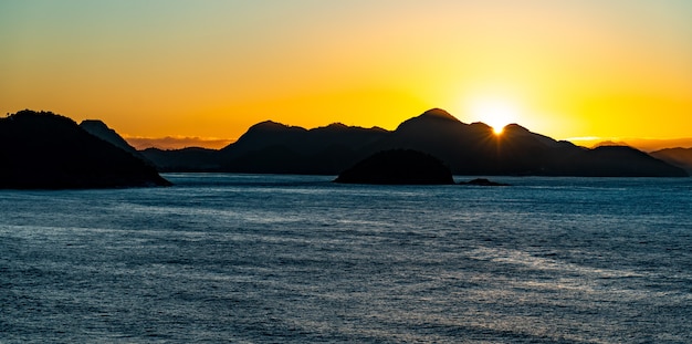 Silhuetas de colinas e rochas à beira-mar durante o pôr do sol no Brasil