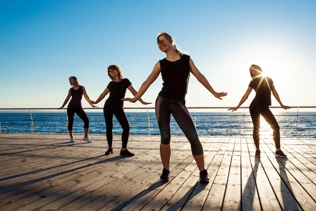 Silhuetas de mulheres esportivas dançando zumba perto do mar ao nascer do sol