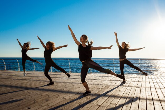 Silhuetas de mulheres esportivas dançando zumba perto do mar ao nascer do sol