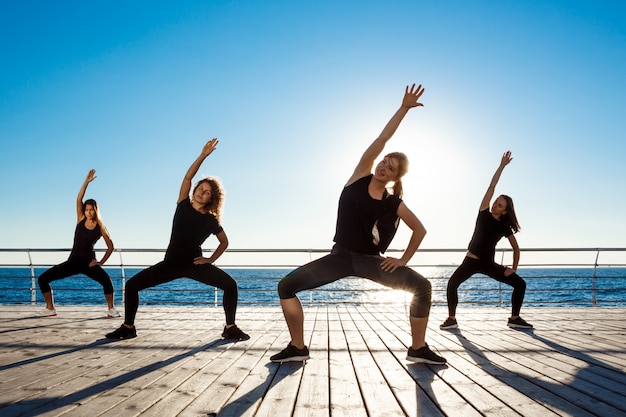 Silhuetas de mulheres esportivas dançando zumba perto do mar ao nascer do sol