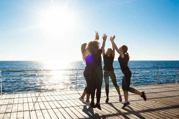 Silhuetas de mulheres esportivas dando highfive perto do mar ao nascer do sol