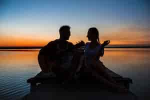 Foto grátis silhuetas do jovem casal lindo descansando regozijando-se ao nascer do sol perto do lago