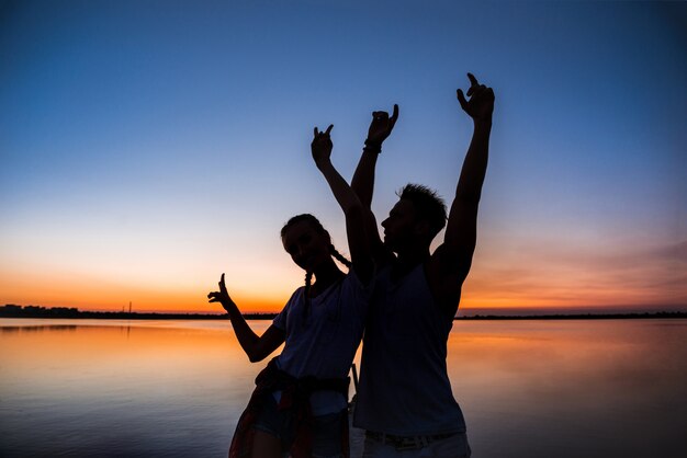 Silhuetas do jovem casal lindo descansando regozijando-se ao nascer do sol perto do lago