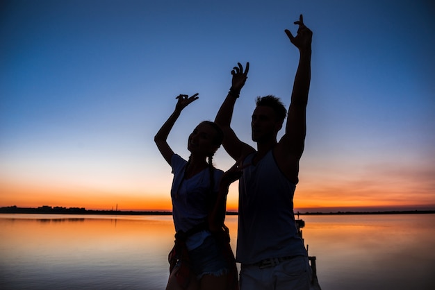 Silhuetas do jovem casal lindo descansando regozijando-se ao nascer do sol perto do lago