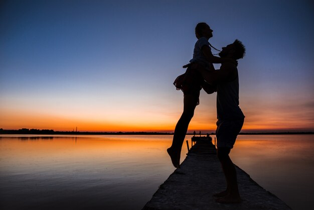 Silhuetas do jovem casal lindo descansando regozijando-se ao nascer do sol perto do lago